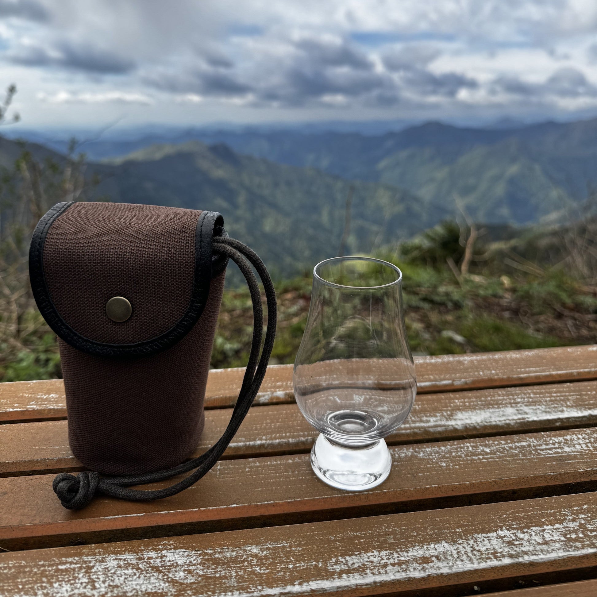 山頂のベンチに置かれた帆布ケースとウイスキーグラス。遠くに山並みが続く